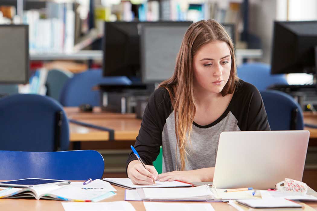 Student sat at a desk in a library making notes while looking at a laptop infront of her